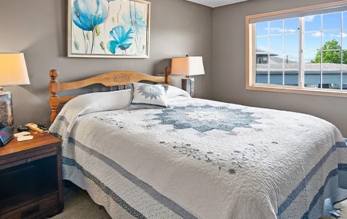 A well-lit bedroom featuring a neatly made bed with a patterned quilt, a bedside lamp, and artwork on the wall.