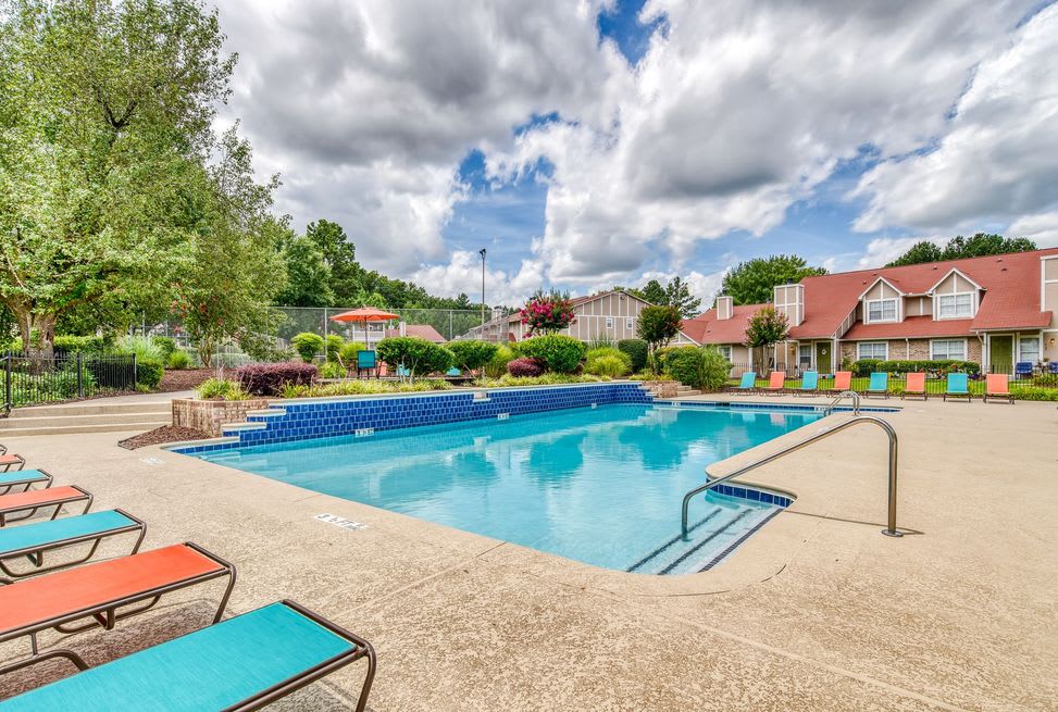 A serene outdoor swimming pool area with lounge chairs under a partly cloudy sky, surrounded by greenery and resort buildings.