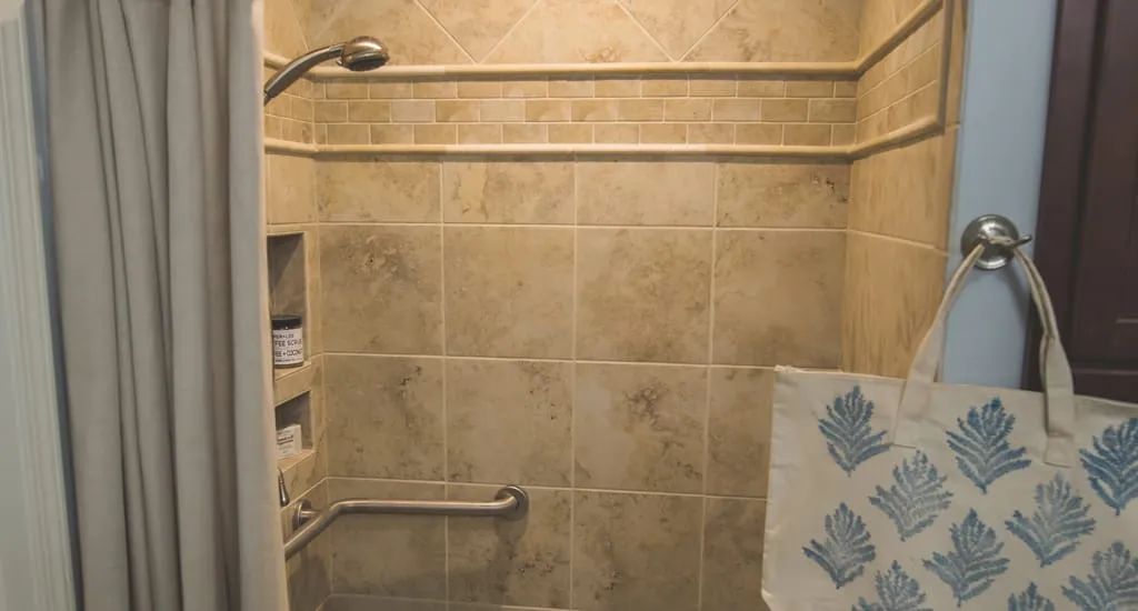 A bathroom shower with beige tiles, a showerhead, a grab bar, and neatly folded towels.