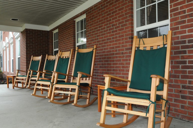 A row of wooden rocking chairs with green cushions on a porch of a brick building.
