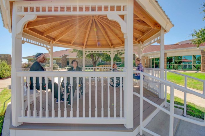 A group of people sitting and conversing inside a wooden gazebo on a sunny day.