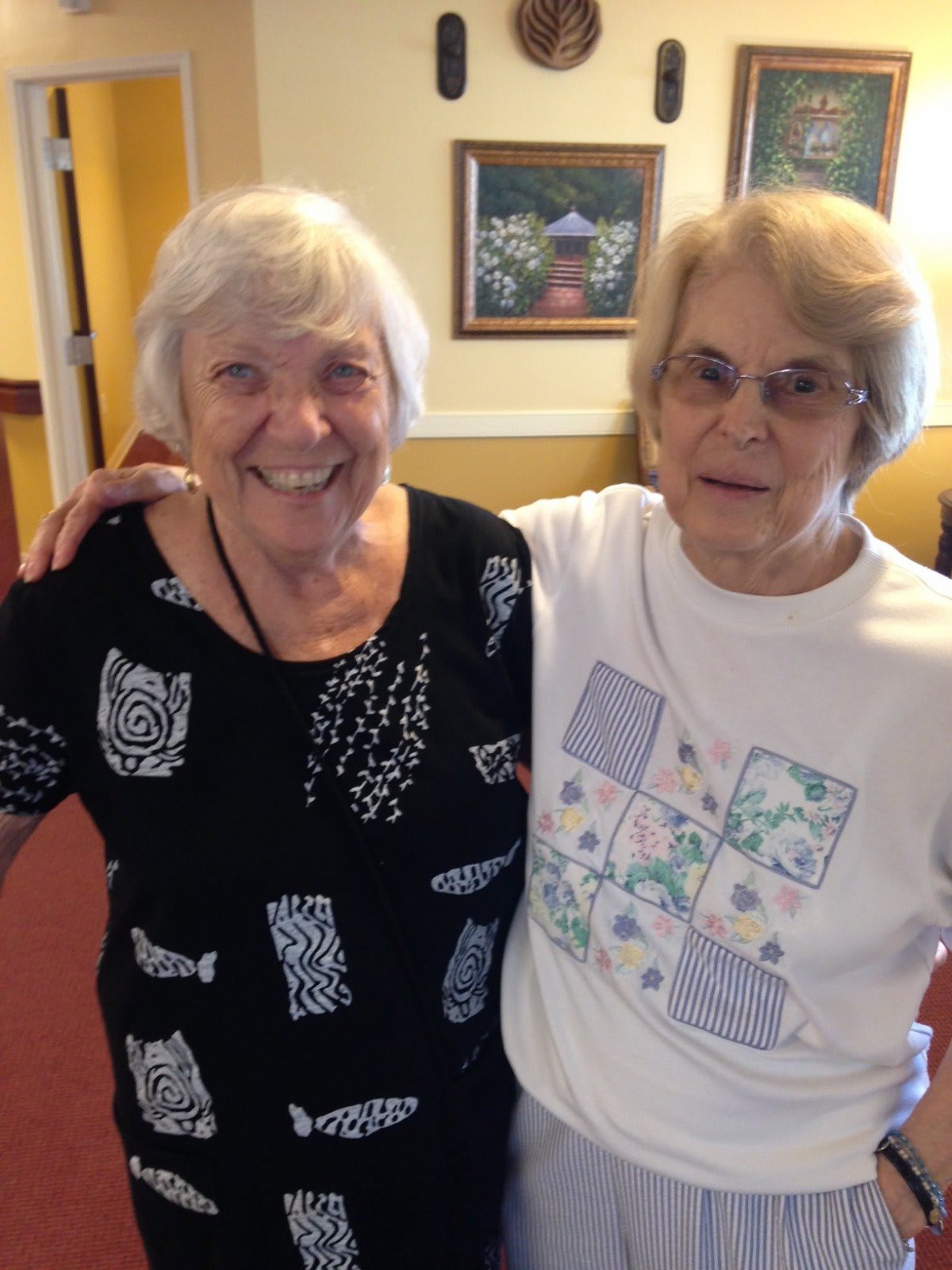 Two elderly women smiling and standing together indoors.
