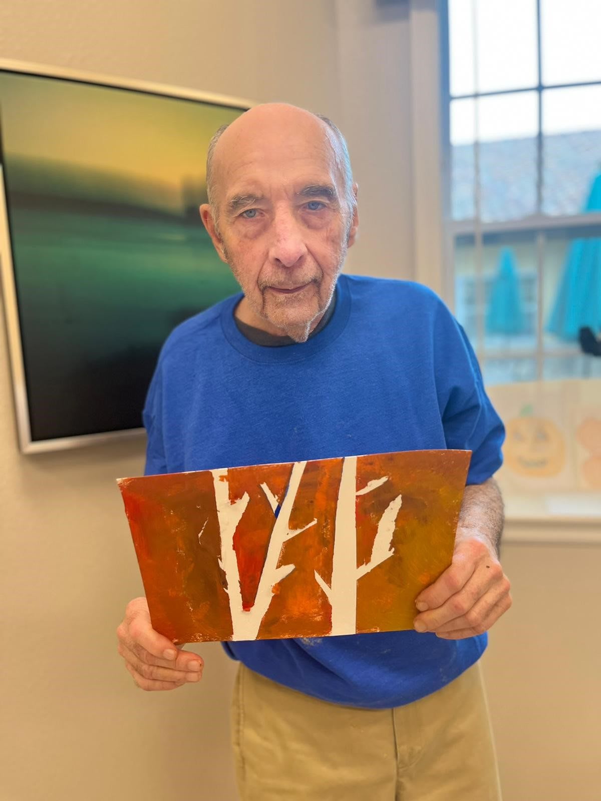 An elderly man in a blue shirt holding a colorful painting of trees indoors.