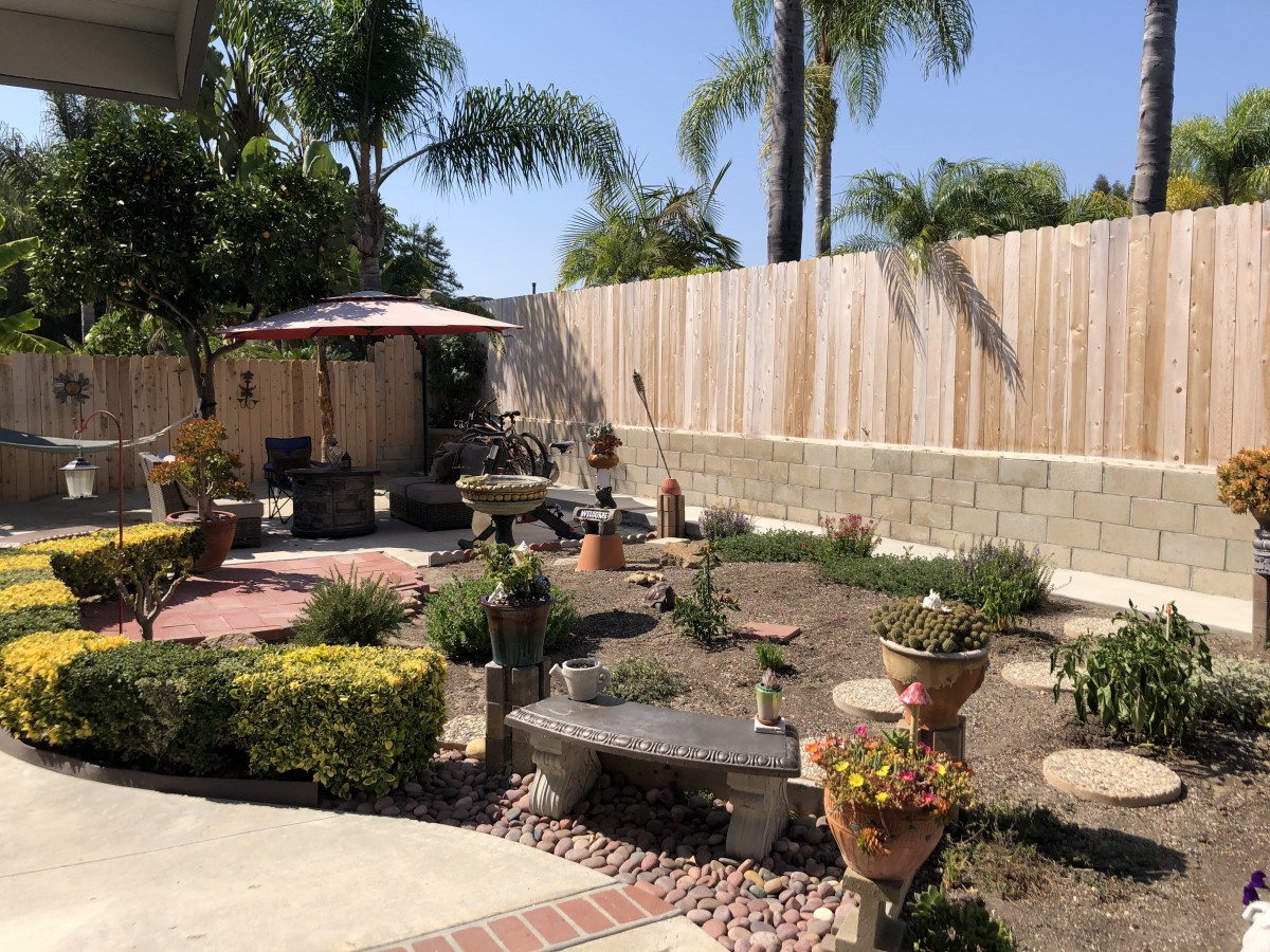 A beautifully landscaped backyard garden with a variety of plants, patio furniture, and a wooden fence under a clear blue sky.