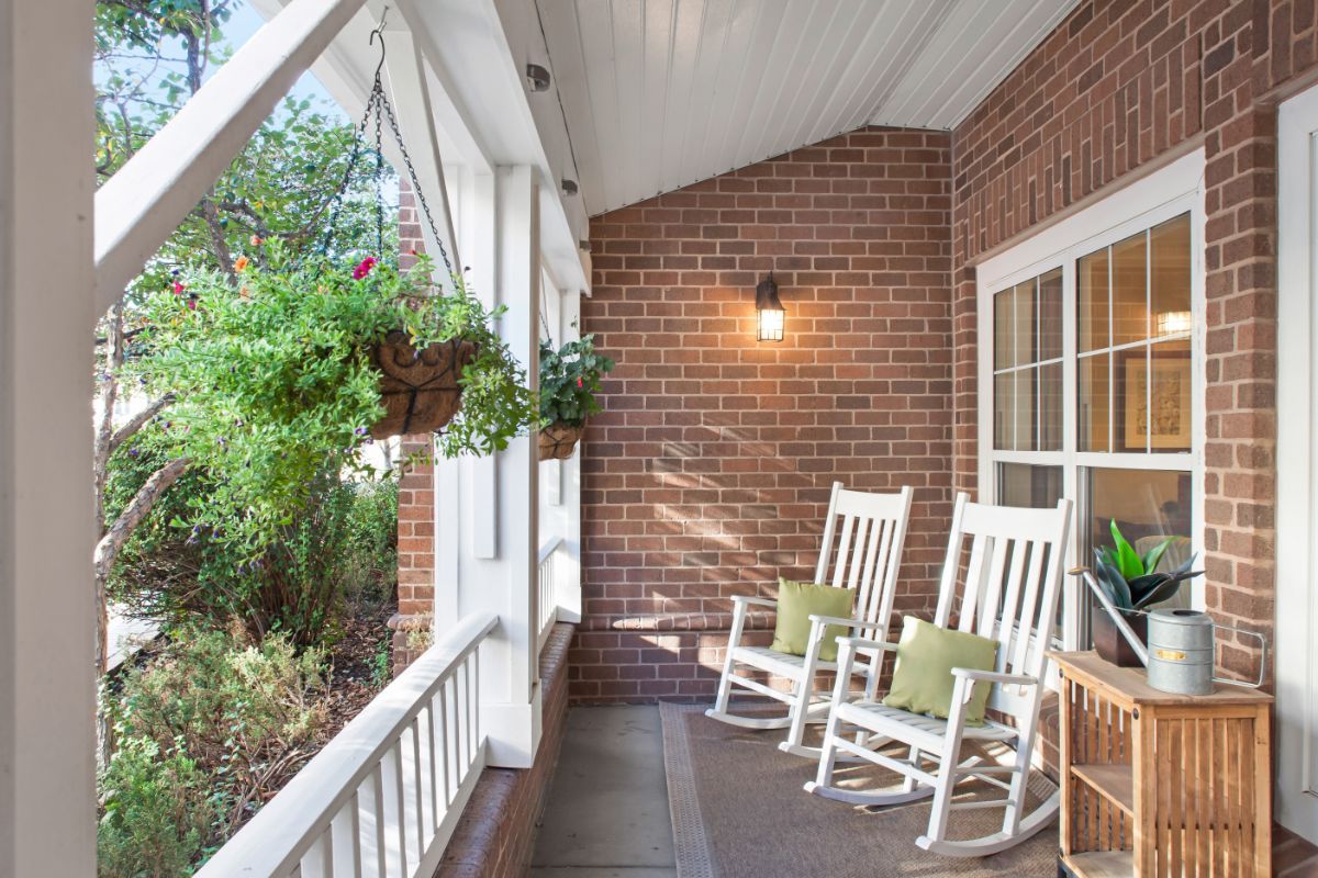 A cozy front porch with white rocking chairs, surrounded by potted and hanging plants against a brick wall.
