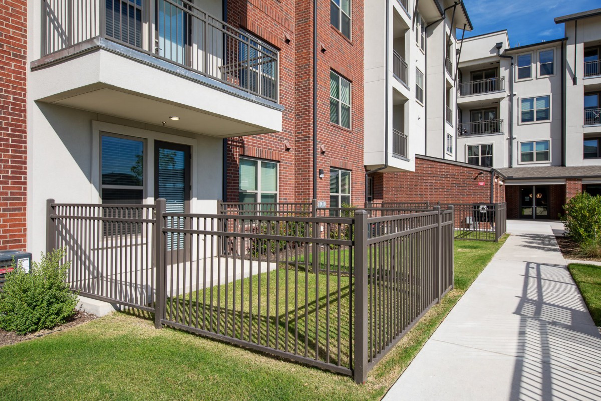 A modern apartment building with a brick exterior, featuring balconies and a fenced lawn area next to the sidewalk.