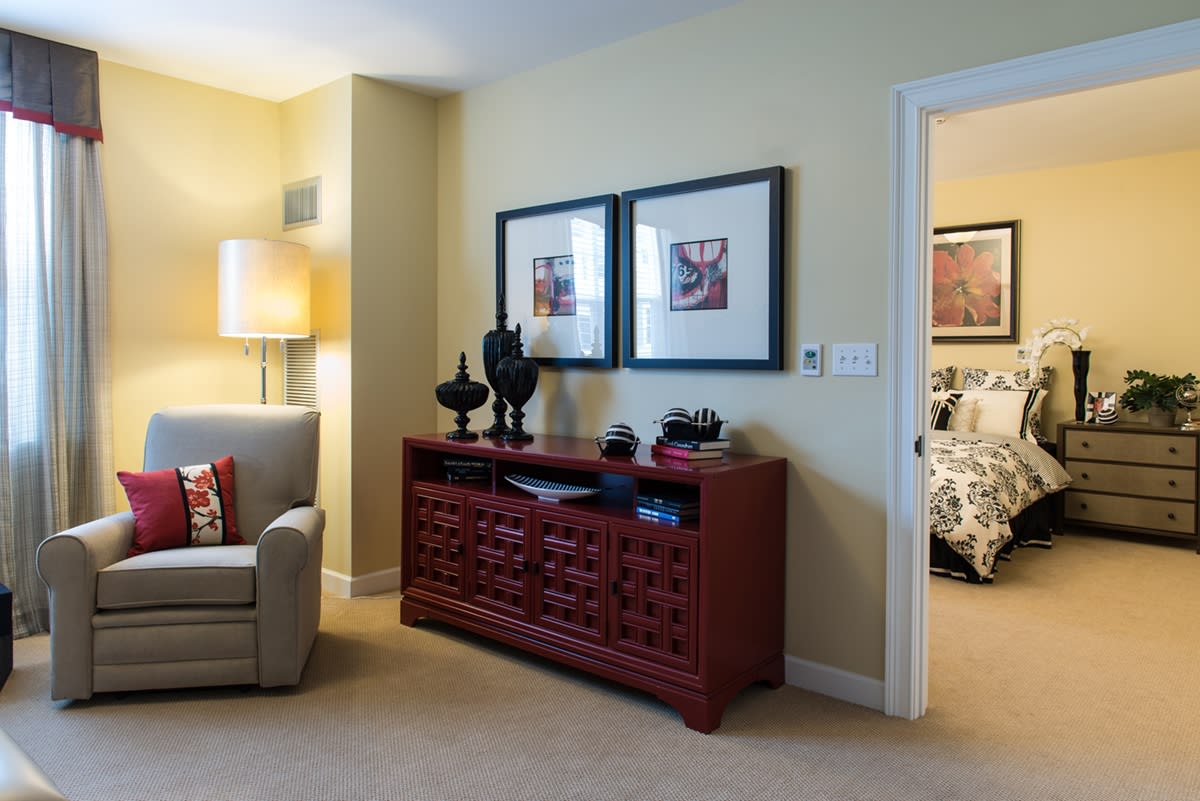 A cozy living room with a comfortable armchair, decorative artwork above a wooden cabinet, and a soft glowing lamp.