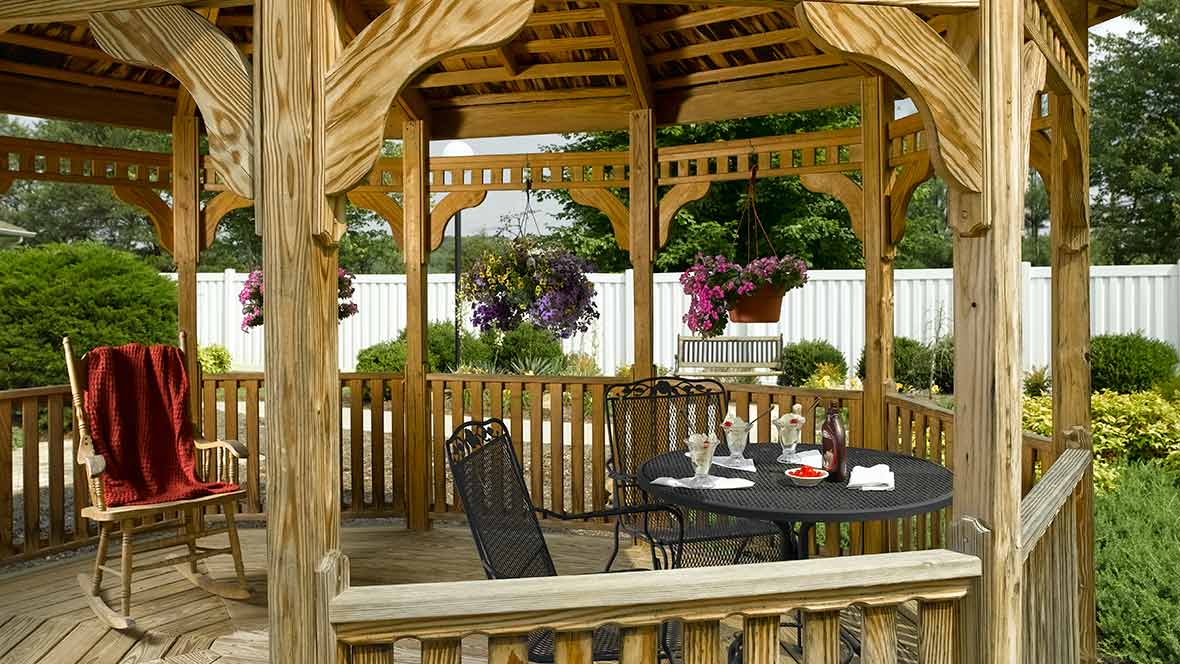 A cozy wooden gazebo with a table and chairs, surrounded by hanging plants in a serene garden setting.