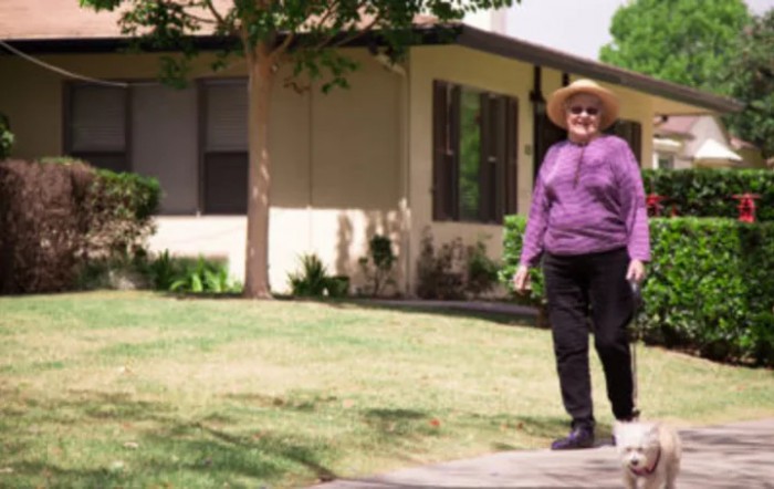 A woman wearing a purple top and hat walks outside a house on a sunny day.