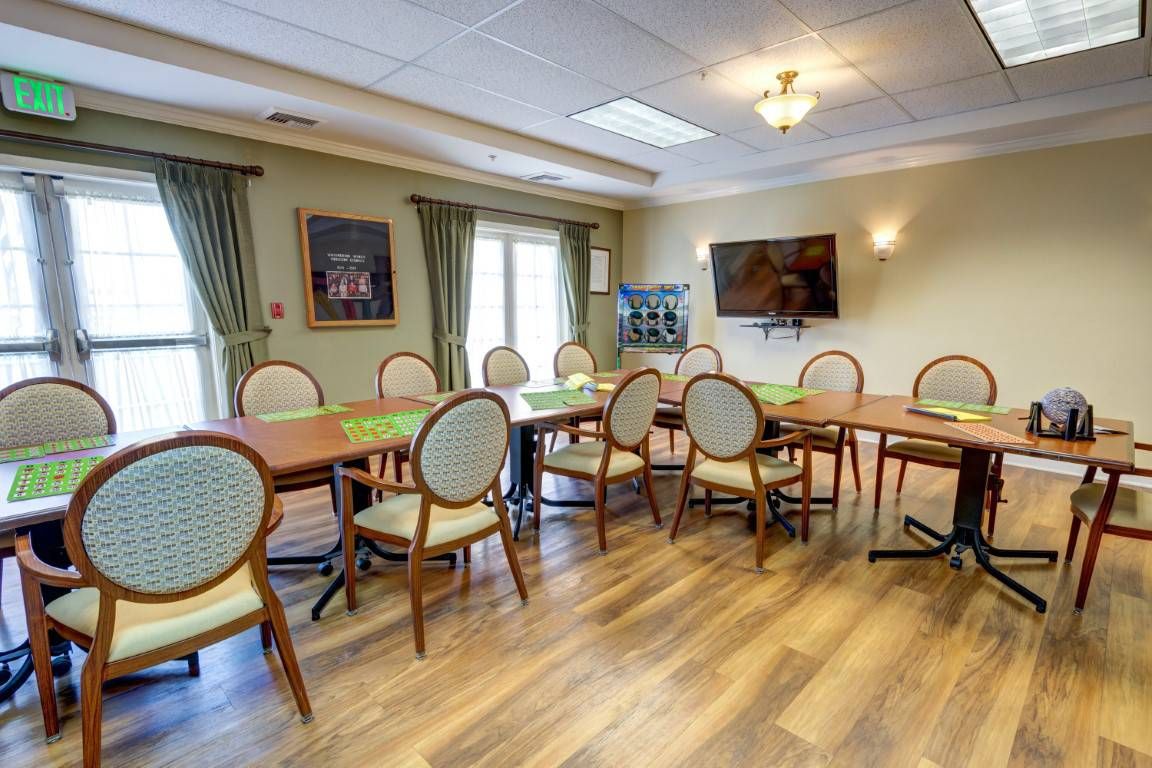 A dining room with tables and chairs arranged on a wooden floor, featuring curtains and wall art.