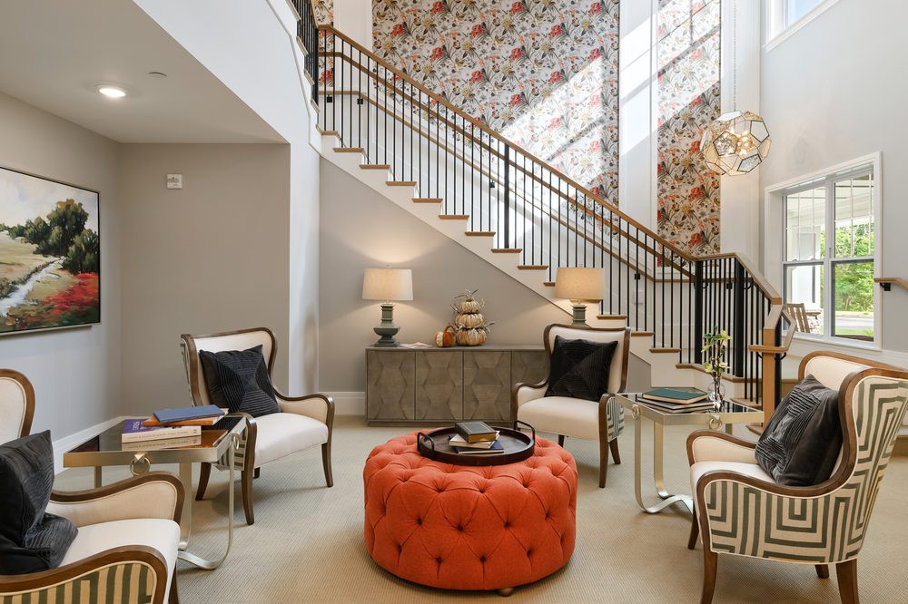 A stylish living room featuring a vibrant orange ottoman, elegant armchairs, and a decorative staircase with floral wallpaper.