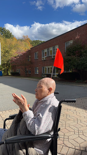 An elderly person in a wheelchair enjoying the outdoors, with a red flag visible against a backdrop of autumn foliage and a building.