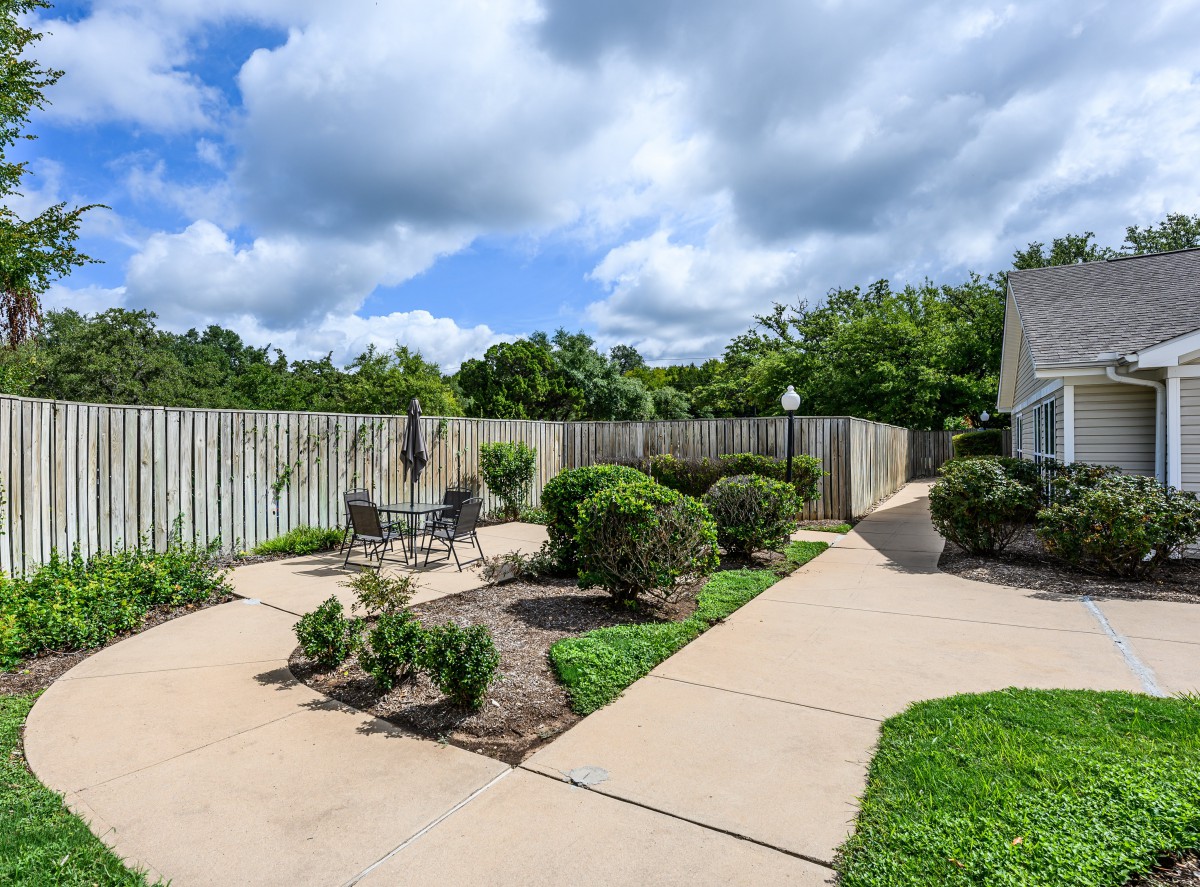 A well-maintained backyard featuring a garden with bushes, a paved patio, and a wooden fence under a partly cloudy sky.