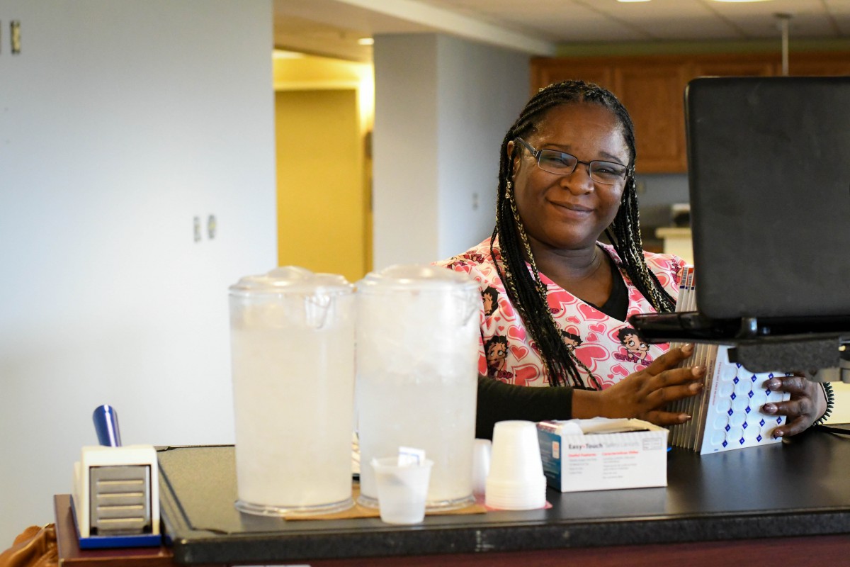 A smiling woman wearing glasses sits at a desk in an office setting.