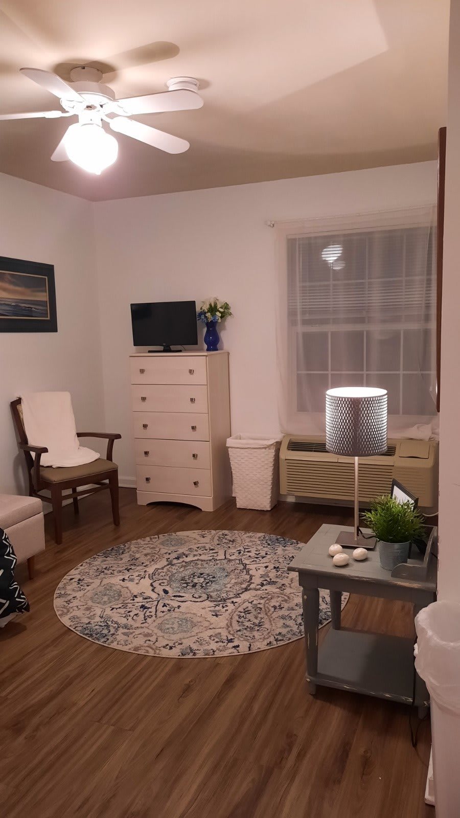 A cozy living room with furniture, including a chair, dresser, and TV, complemented by a decorative rug and lamp.