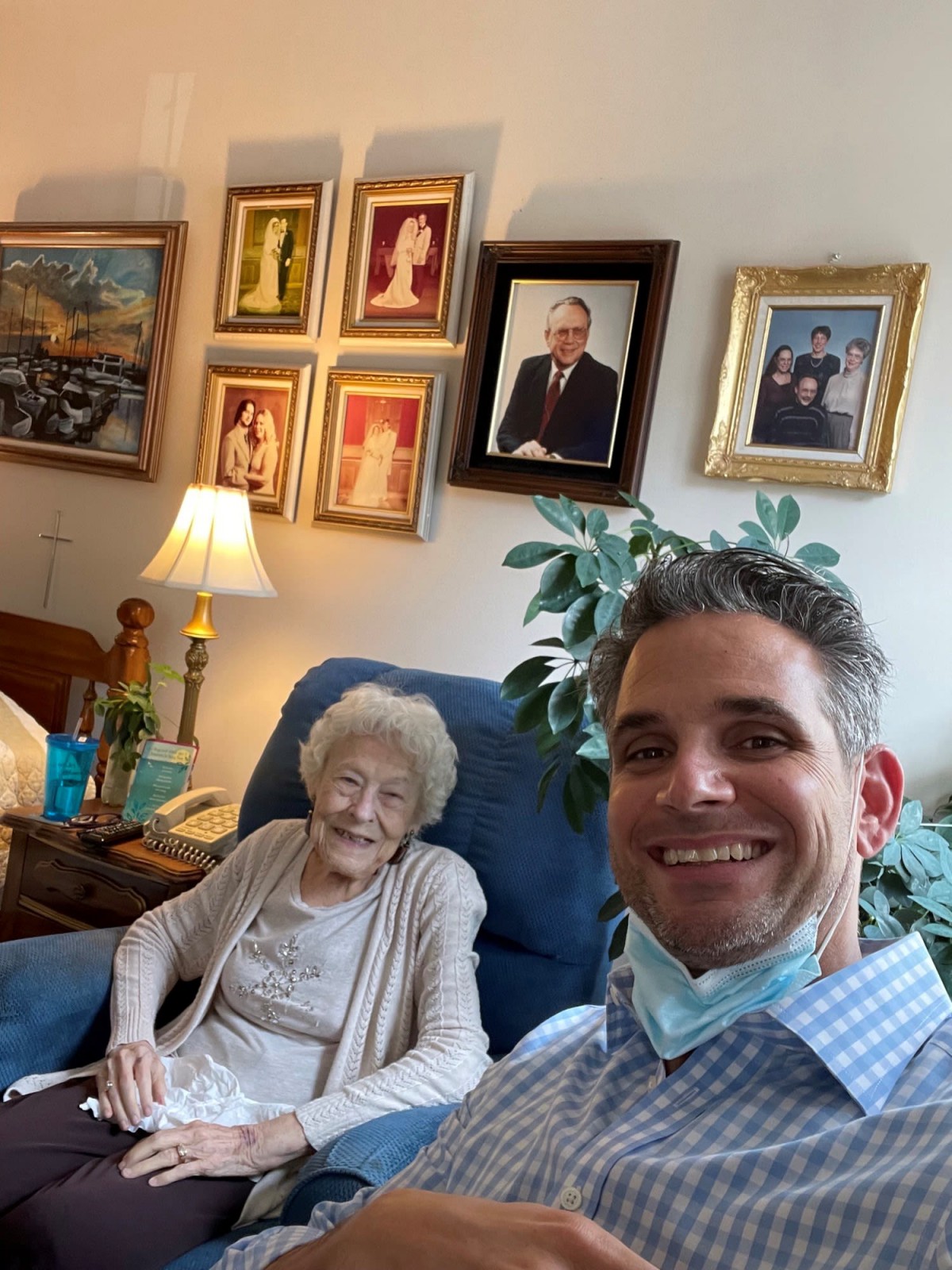 A man takes a selfie with an elderly woman seated in a living room filled with framed pictures on the wall.