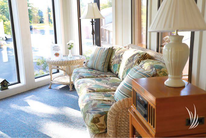 A sunlit room with a floral-patterned sofa and cushions next to large windows, featuring a lamp and a small table.
