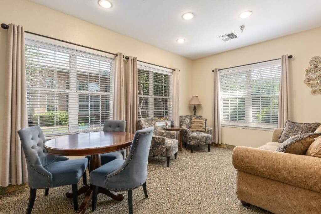 A cozy living room with elegant furnishings, featuring a table and chairs, a sofa, and large windows with curtains.