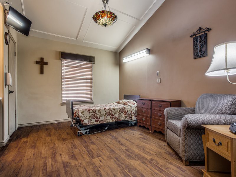A hospital room with a single bed, wooden dresser, and armchair, featuring a cross on the wall and a window with blinds.