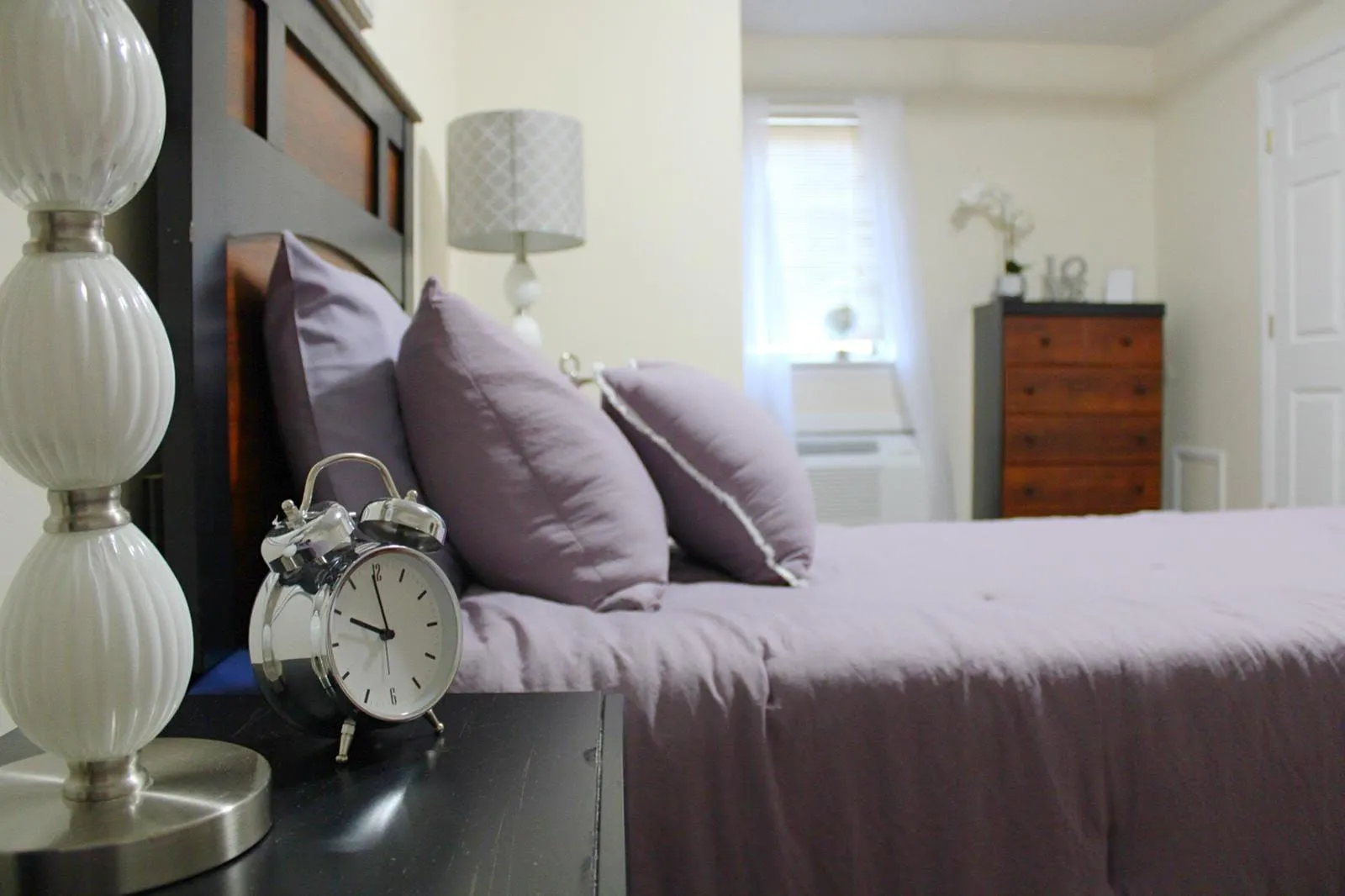 A cozy bedroom with a neatly made bed, alarm clock on the side table, and soft lighting.