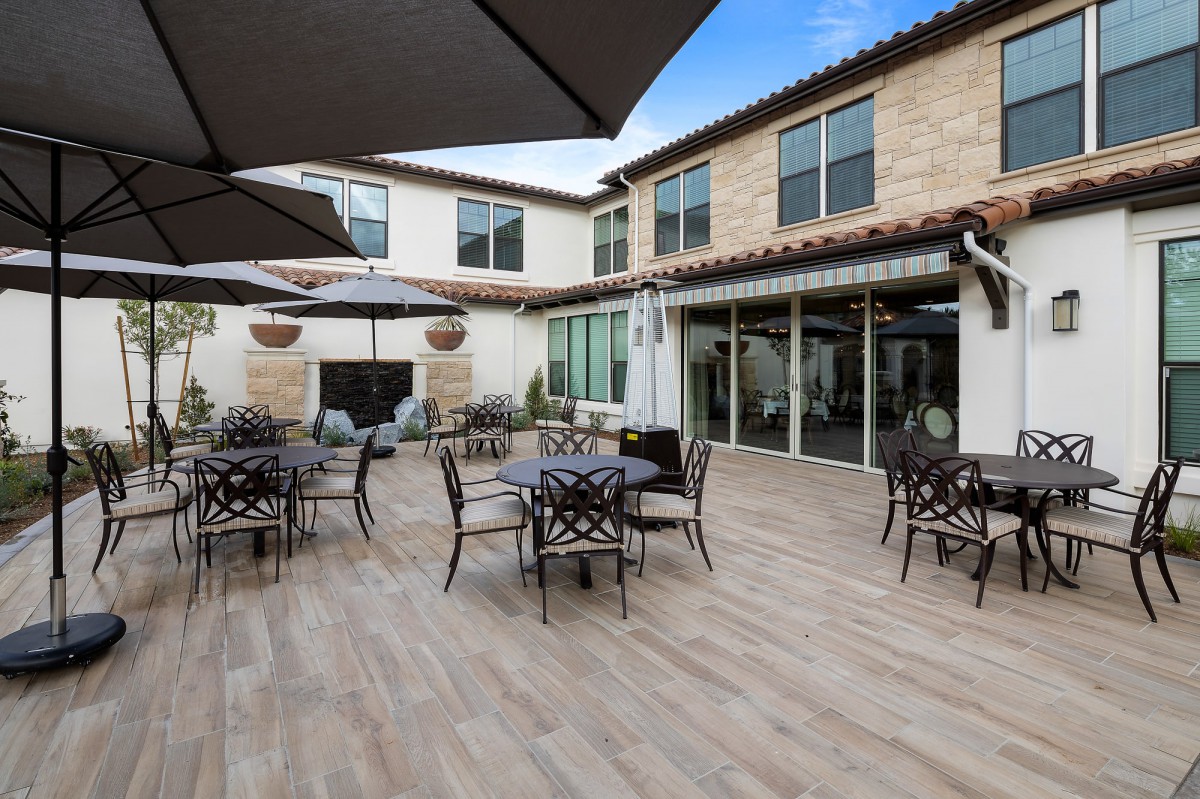 A spacious outdoor patio area with tables, chairs, and umbrellas in front of a modern building with a stone facade.