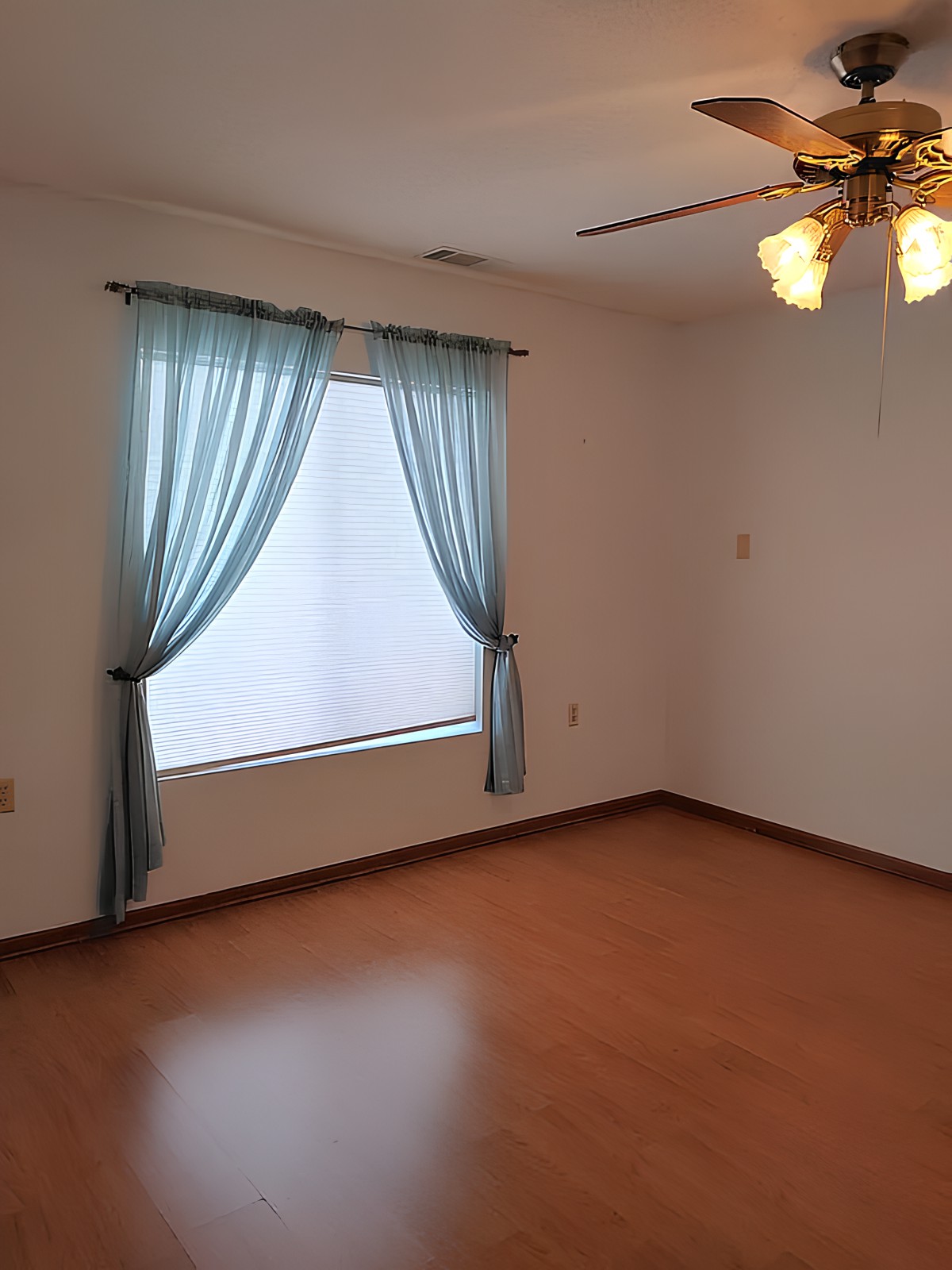 An empty room with wooden flooring, featuring a window with blue curtains and a ceiling fan.