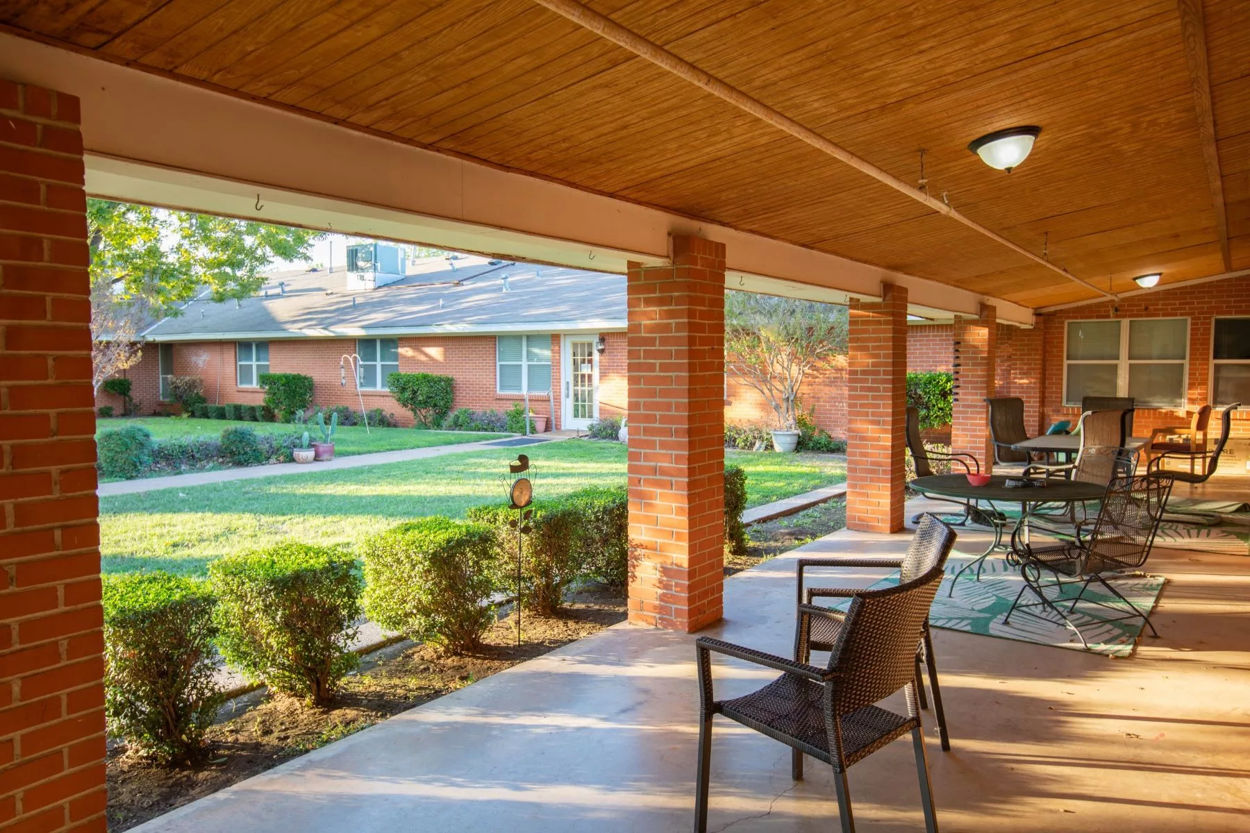 A sunny patio with chairs and a table overlooks a well-maintained garden and brick building.