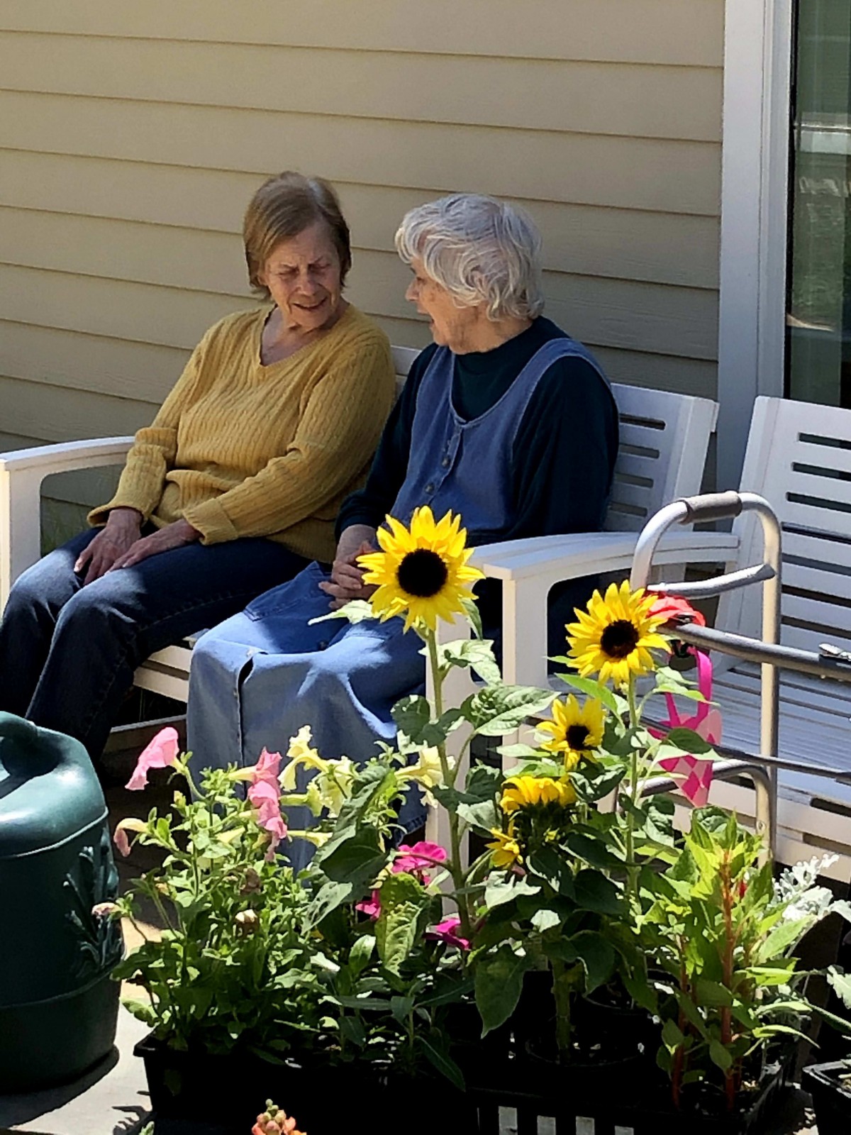 Two elderly women sit and chat on a bench in a garden surrounded by flowers.