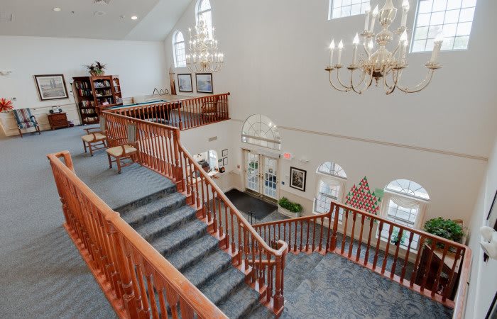 A grand interior with a large wooden staircase, chandeliers, and ample natural light from tall windows.