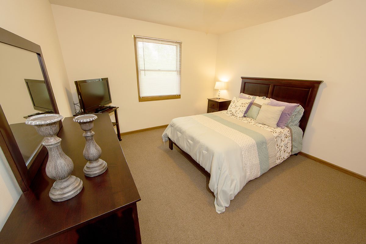 A cozy bedroom featuring a bed with decorative pillows, a dresser with a TV, and a warmly lit lamp.