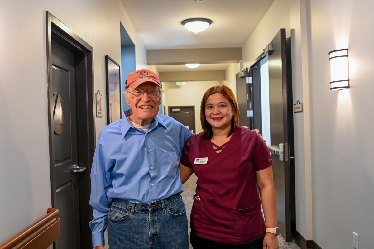 A friendly nurse with a senior man in a hallway, both smiling.