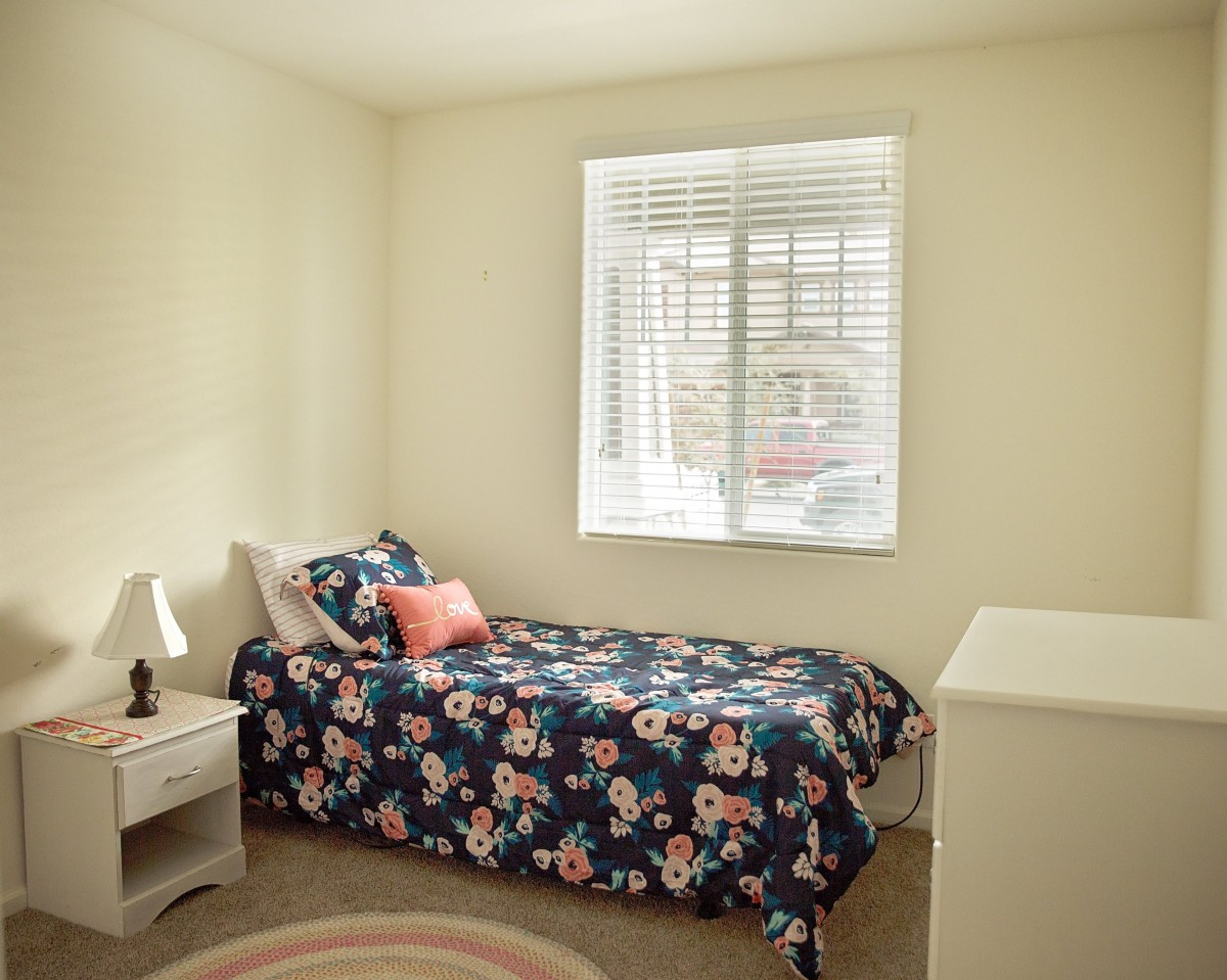 A cozy bedroom with a single bed featuring floral bedding, a small lamp on the nightstand, and a window letting in natural light.