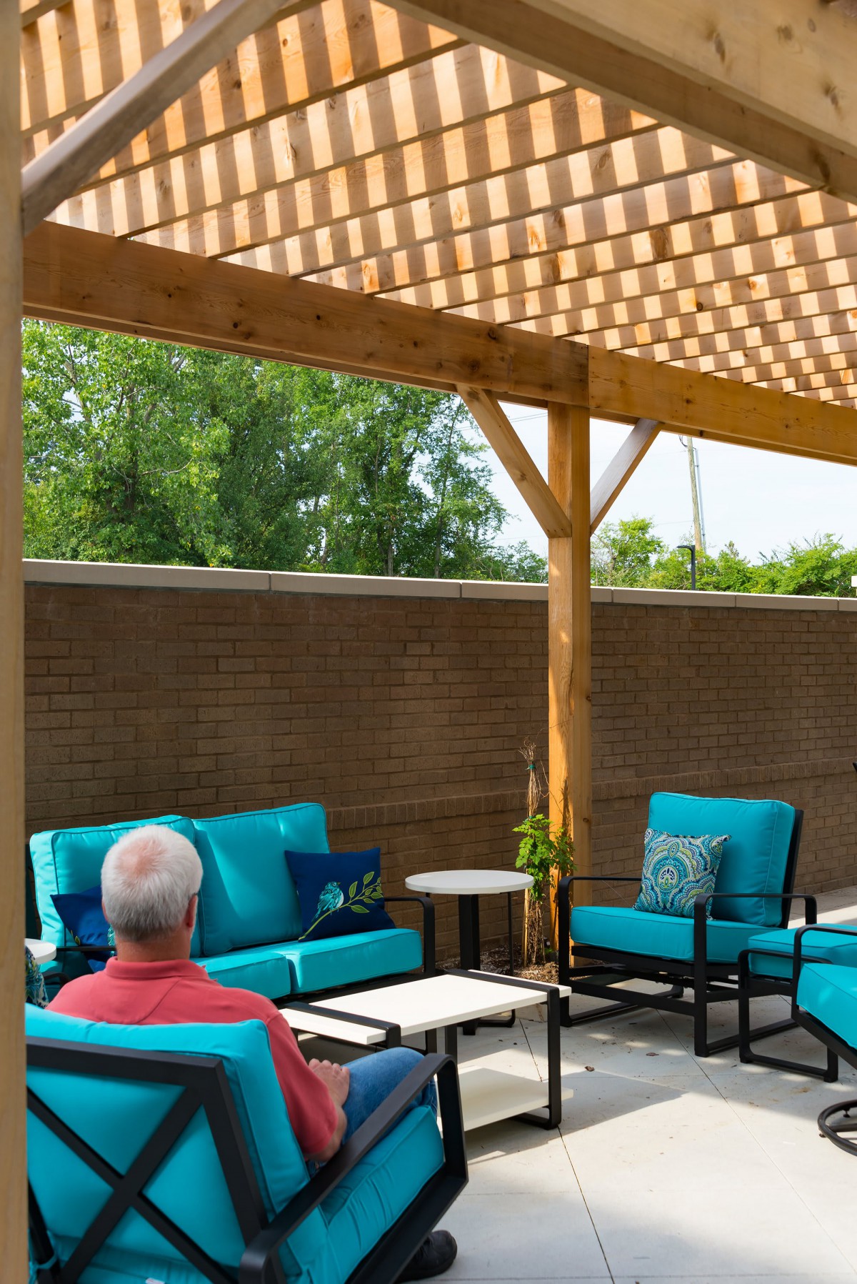 A man relaxes under a wooden pergola with teal outdoor furniture in a backyard setting.