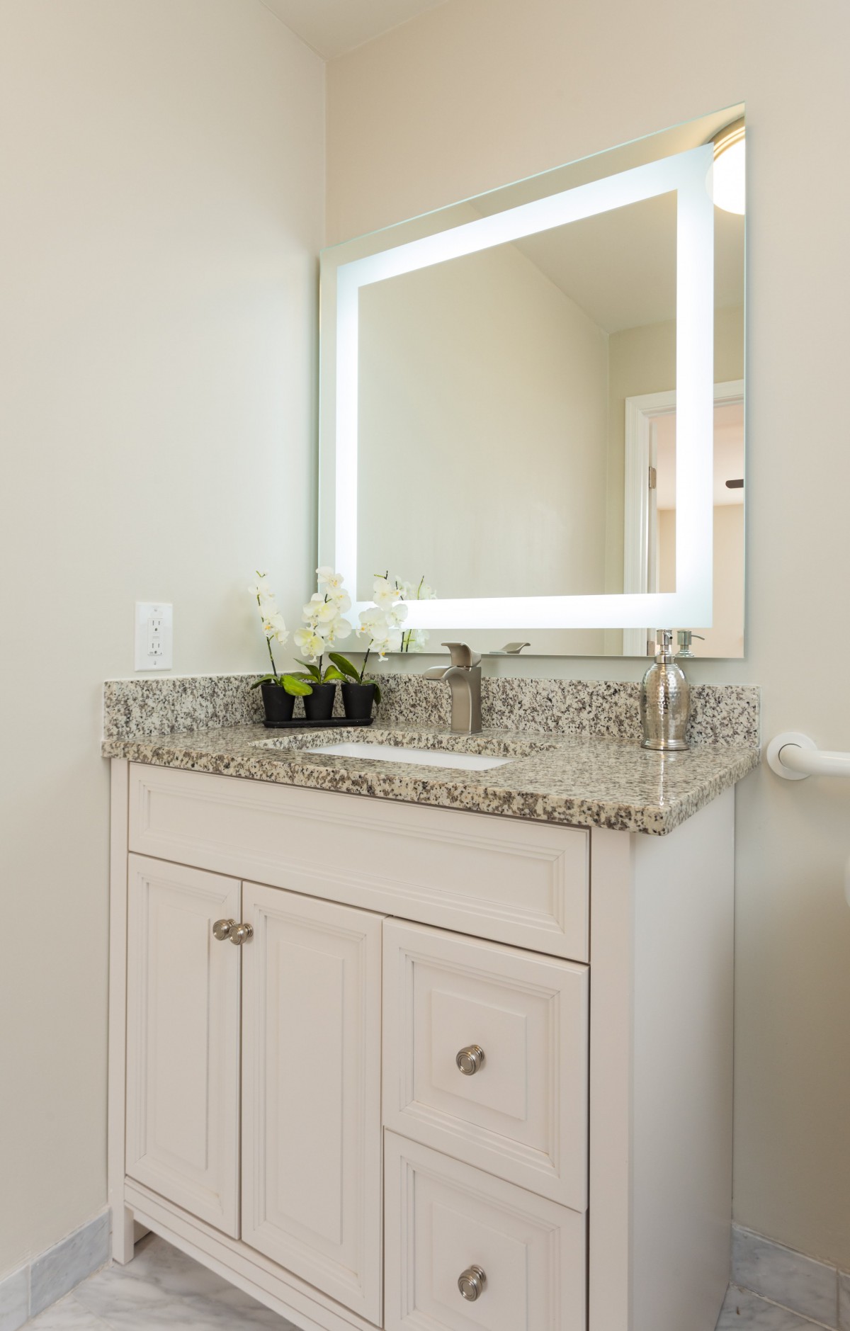 A modern bathroom vanity with a granite countertop, illuminated mirror, and decorative flowers.
