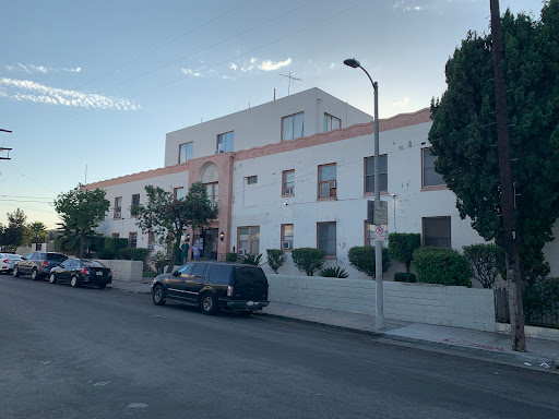 A view of a residential building along a street, with parked vehicles and green shrubbery.