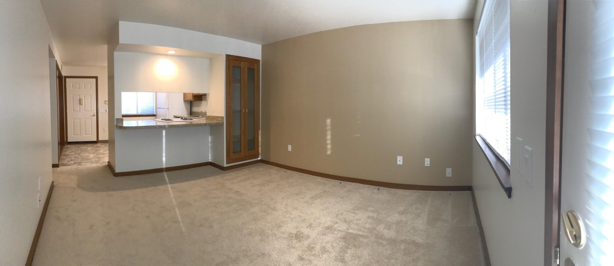 An empty room with beige walls and carpet flooring, featuring a view into a kitchen area.
