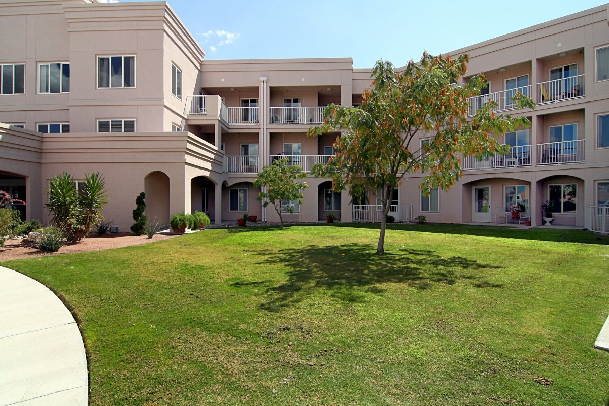 A sunny view of a three-story building with balconies and a well-maintained lawn with a tree in the foreground.