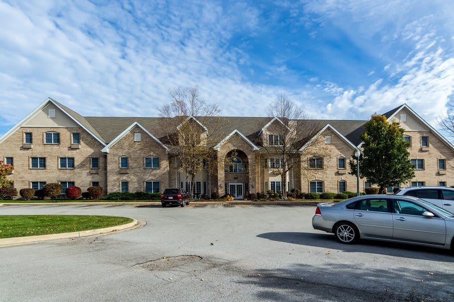 A large residential building with a parking lot in front and cars parked under a clear blue sky with clouds.