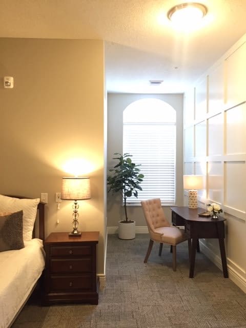 A cozy bedroom with a desk, chair, and decorative plant by a large window.