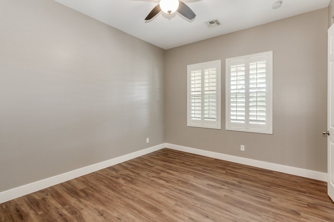 An empty room with hardwood floors, a ceiling fan, and window shutters.