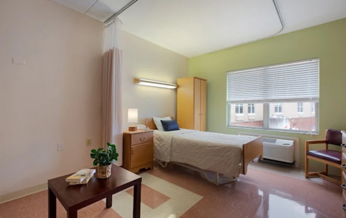 A clean and minimalist hospital room featuring a single bed, wardrobe, and window providing natural light.