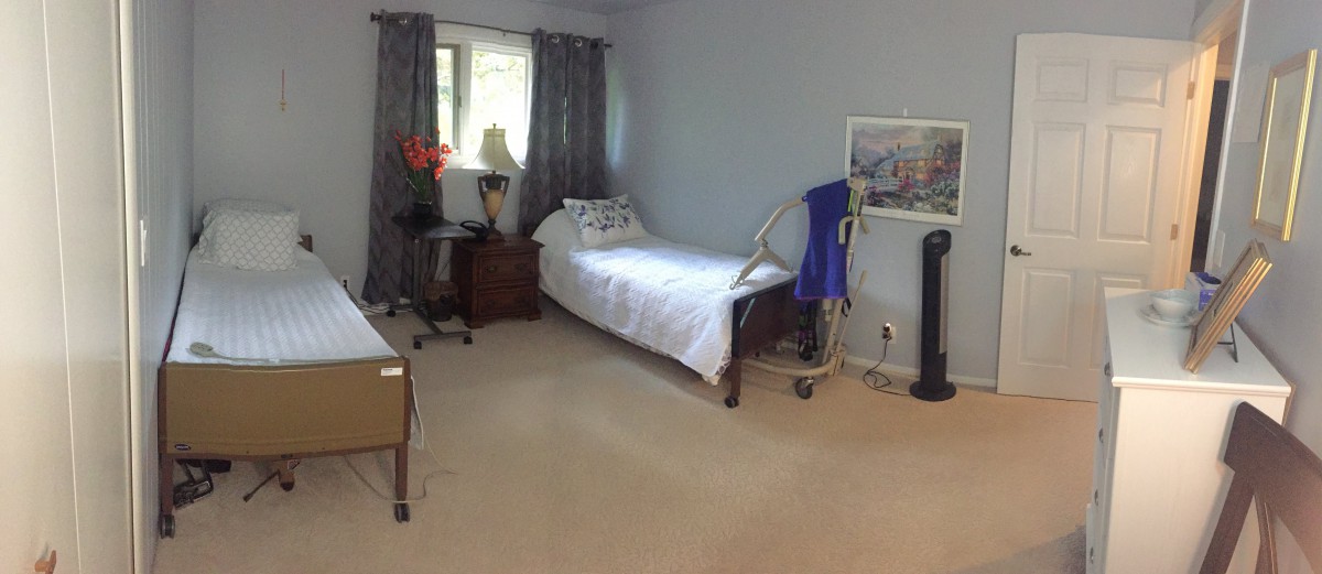 A tidy bedroom featuring a hospital bed, side table with a lamp, and natural light from a window.