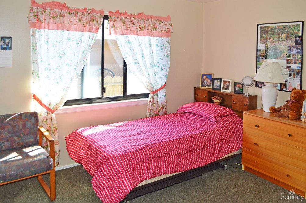 A cozy bedroom with a single bed dressed in pink bedding, complemented by floral curtains and a small wooden nightstand.