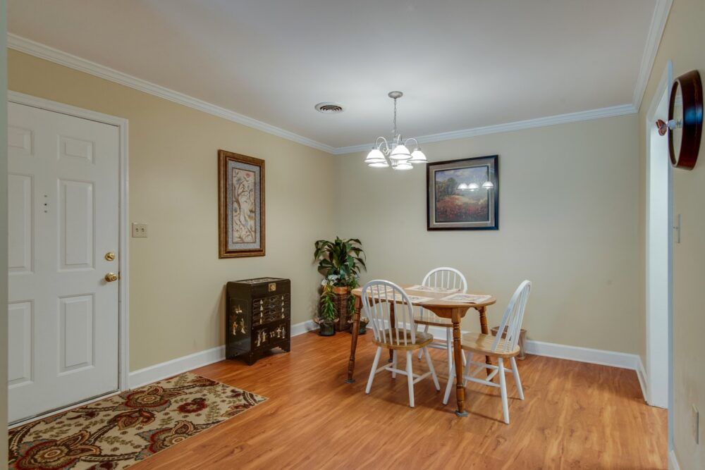 A cozy dining room with a wooden floor, featuring a small table with chairs, decorative artwork on the walls, and a potted plant.