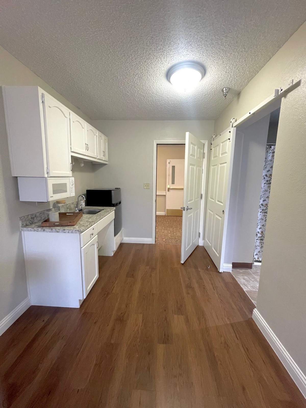 A compact kitchen area with white cabinets and wooden flooring, featuring a microwave and an open door leading to another room.