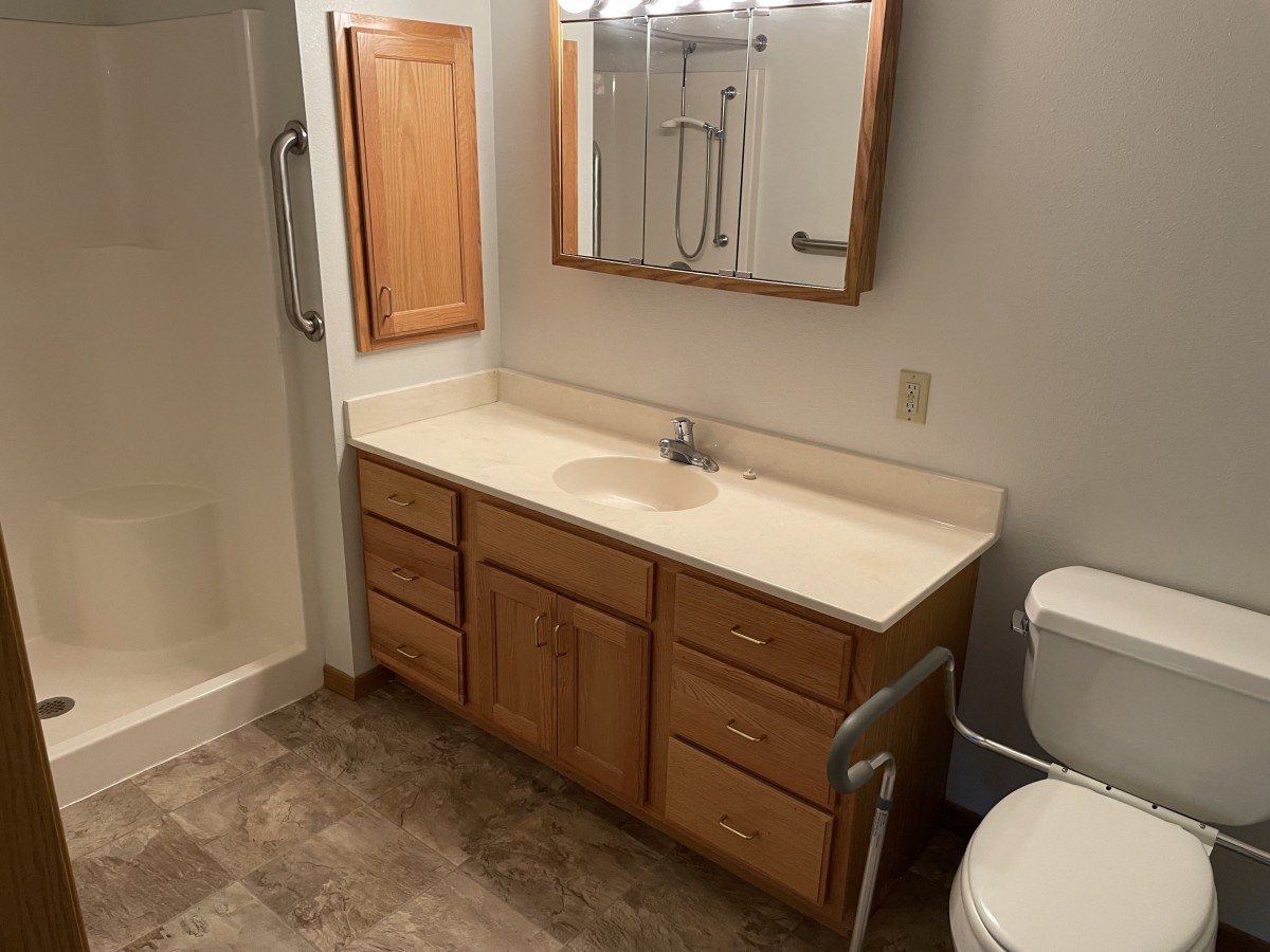 A bathroom with a wooden cabinet, sink, mirror, toilet, and shower featuring a grab bar.