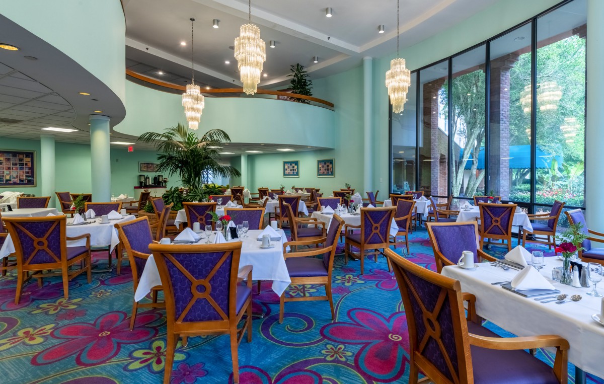 A spacious restaurant interior with elegantly set tables and colorful decor, featuring large chandeliers and a vibrant carpet.
