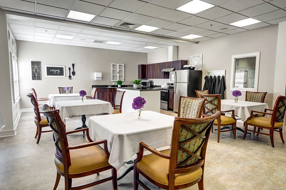 A modern dining area with neatly arranged tables and chairs, adorned with purple flowers, adjacent to a kitchen space.