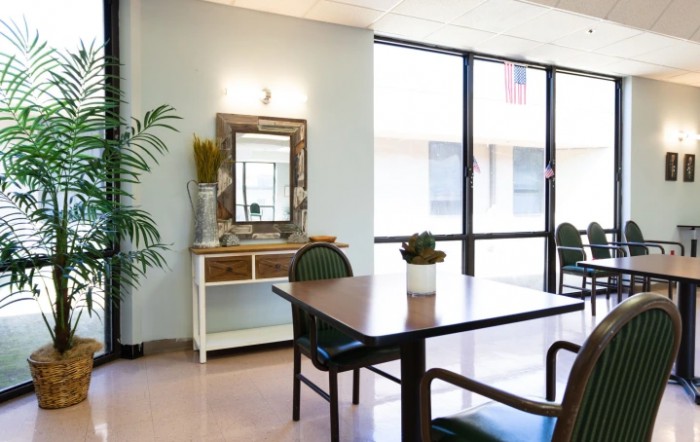 A modern living room with a wooden table, chairs, a decorative mirror, and large windows allowing natural light.
