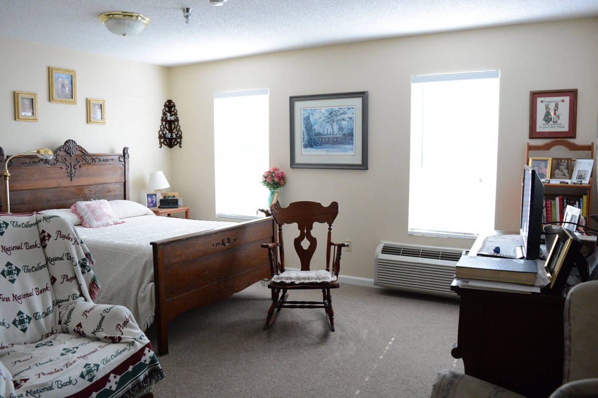 A cozy bedroom with a wooden bed, chair, and two windows bringing in natural light.